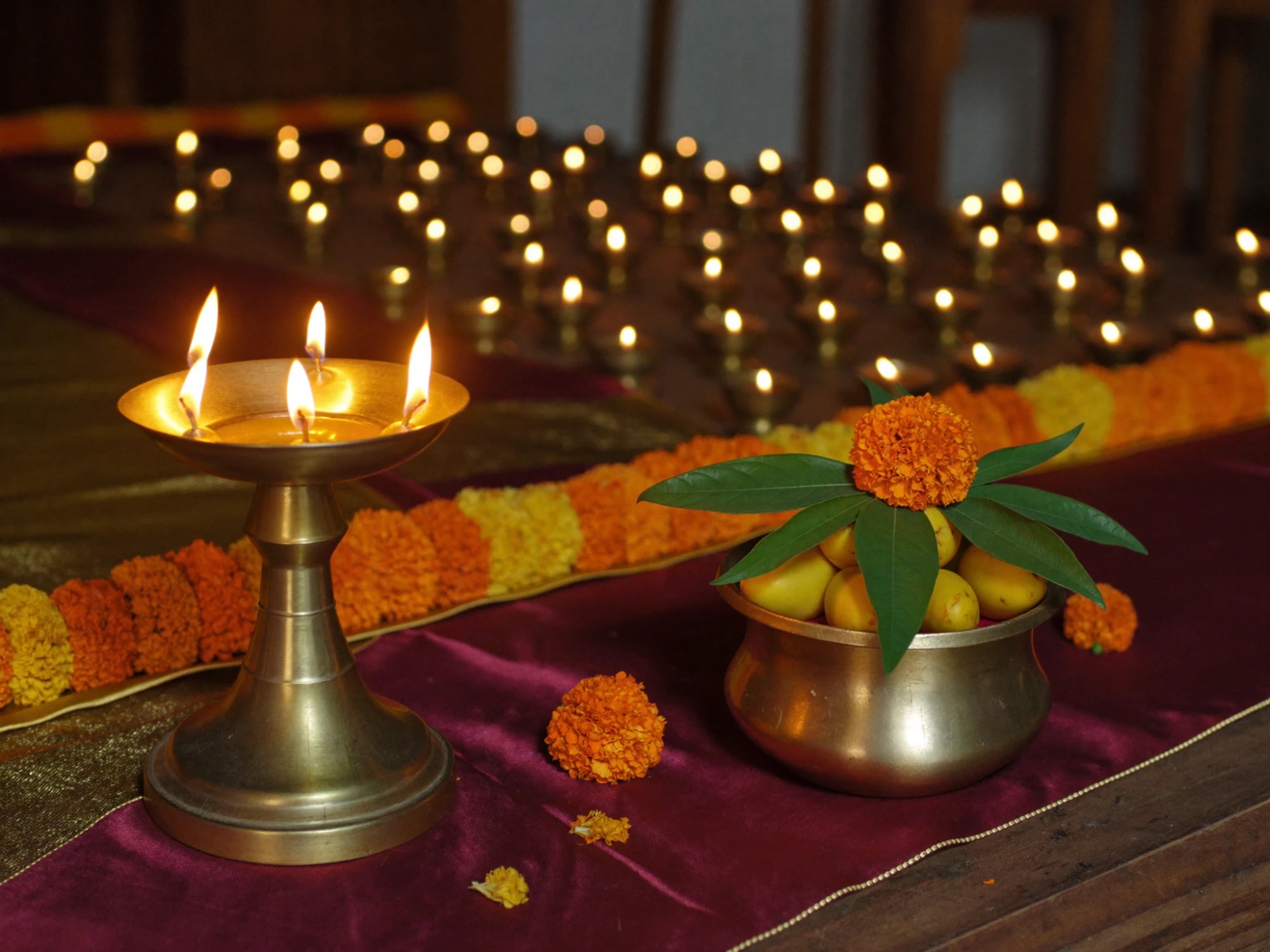 A brass diya and a kalash filled with mangoes and marigold on a maroon cloth, with rows of oil lamps behind, marking an auspicious offering